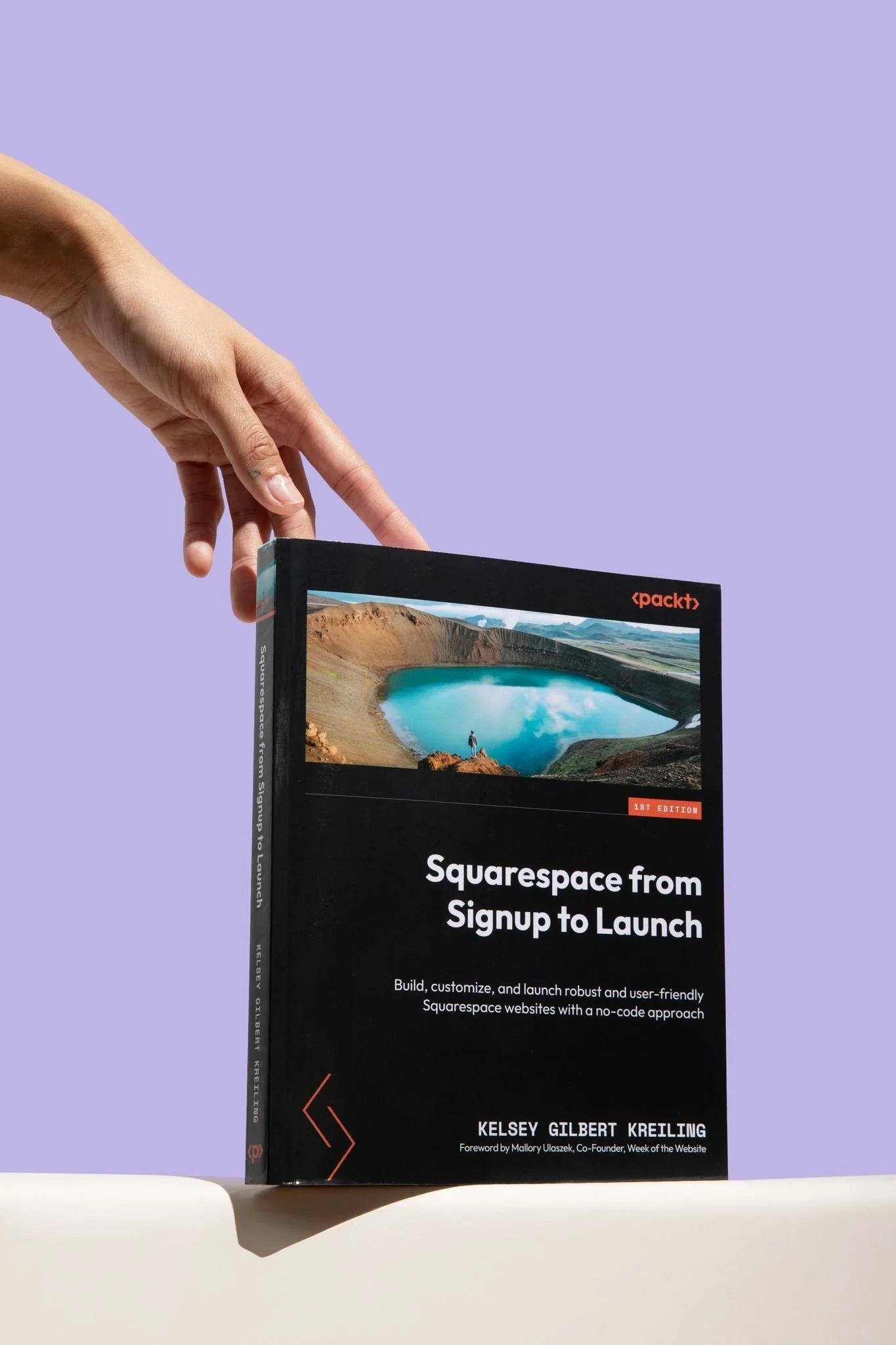 A book titled 'Squarespace from Signup to Launch' by Kelsey Gilbert Kreiling, with a cover photo of a crater lake and a hand pointing at the book, against a purple background.