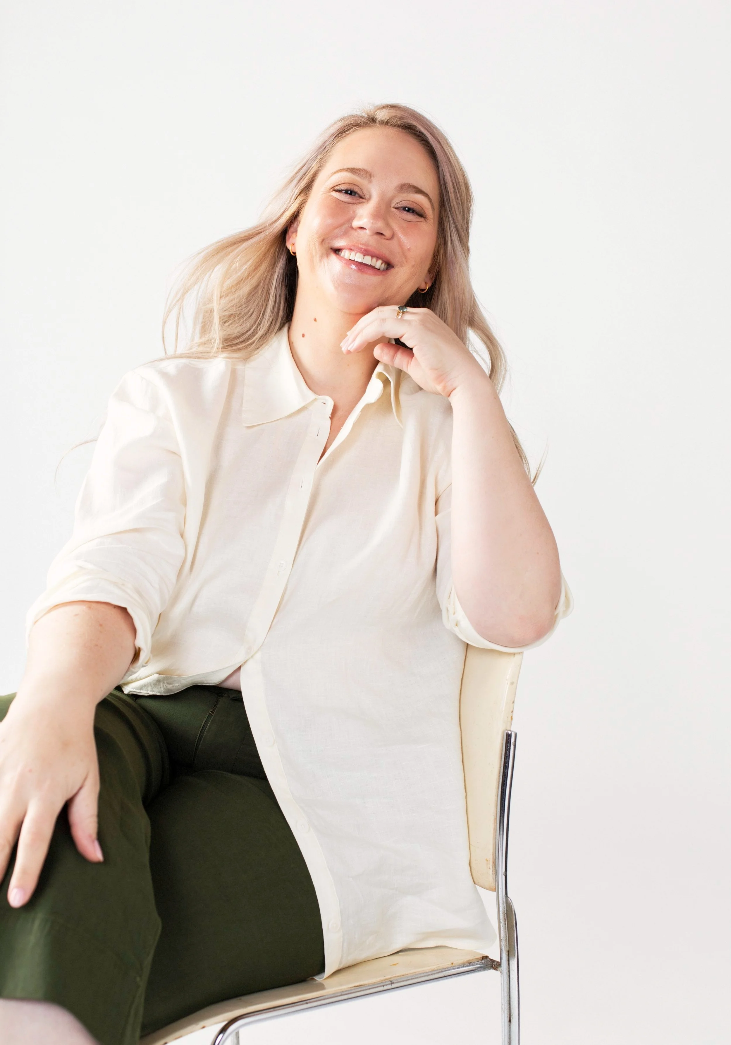 A smiling woman with blonde hair, wearing a white shirt, sitting on a chair, with her hand resting near her chin, leaning slightly towards a person seated nearby, against a plain white background.
