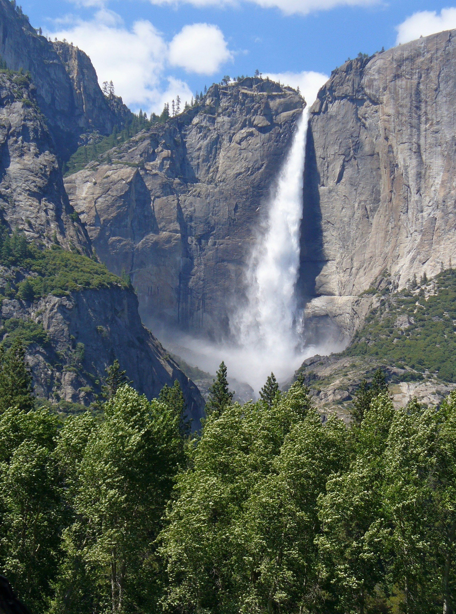 View of Yosemite Falls cascading down granite cliffs, with green trees in the foreground and a partly cloudy sky.