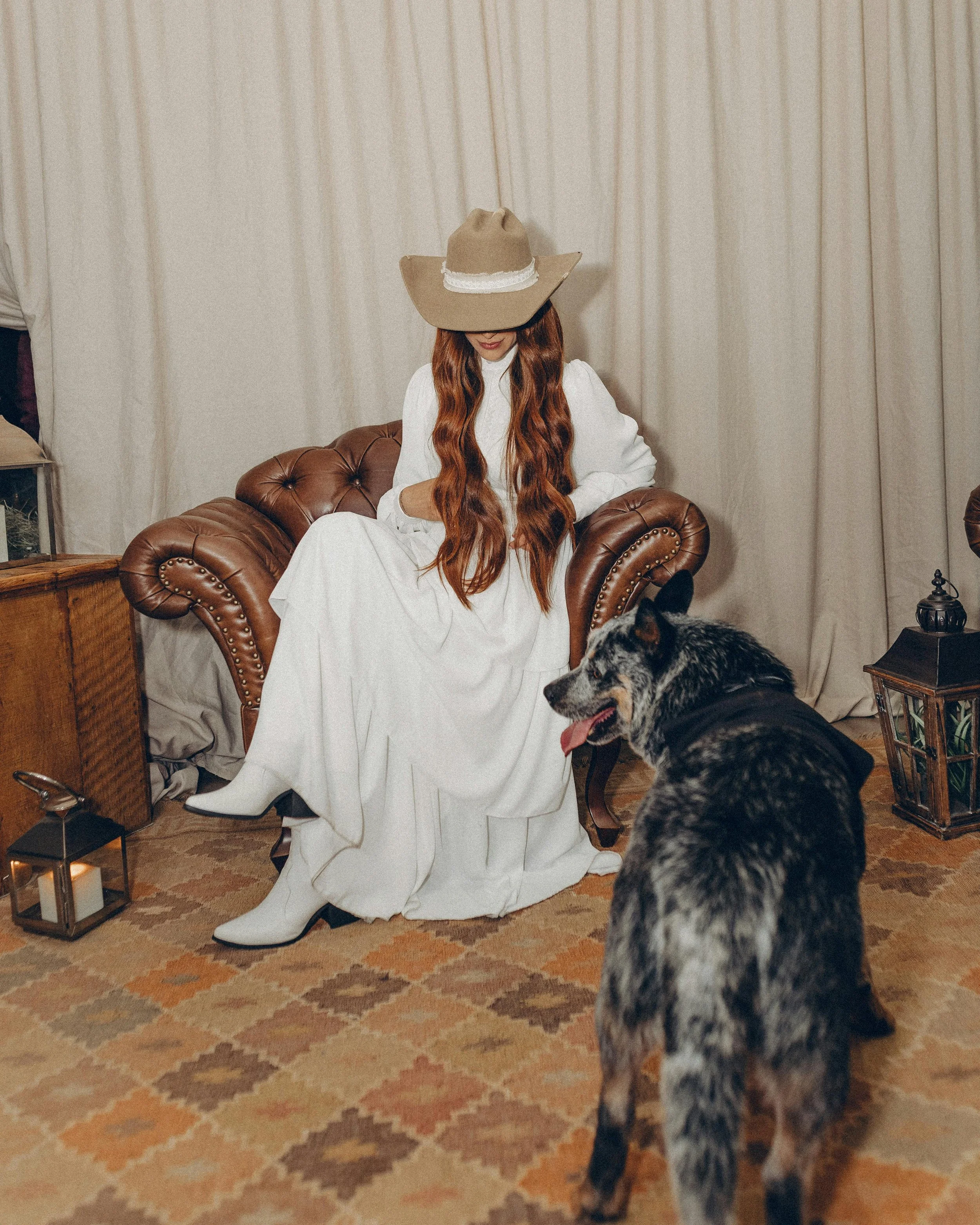 A woman with long red hair sitting on a leather couch, wearing a white dress and a wide-brimmed hat, with a large dog in front of her, inside a room with curtains and vintage-style decor.