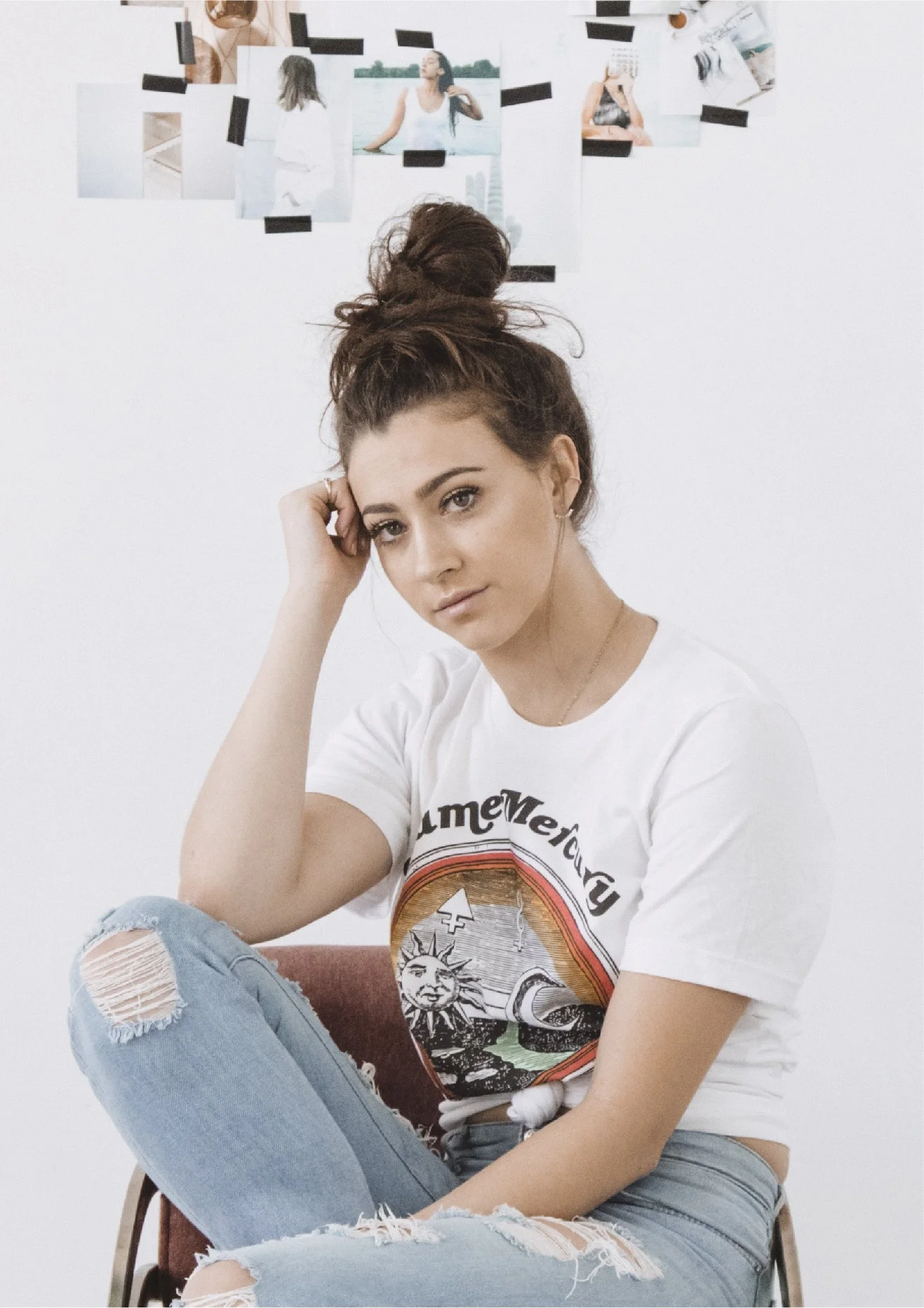 A woman with a top knot hairstyle sitting on a chair with torn jeans, wearing a white graphic t-shirt, looking at the camera with her hand resting on her head. In the background, a white wall with photos taped on it, including images of a woman by water and other scenes.