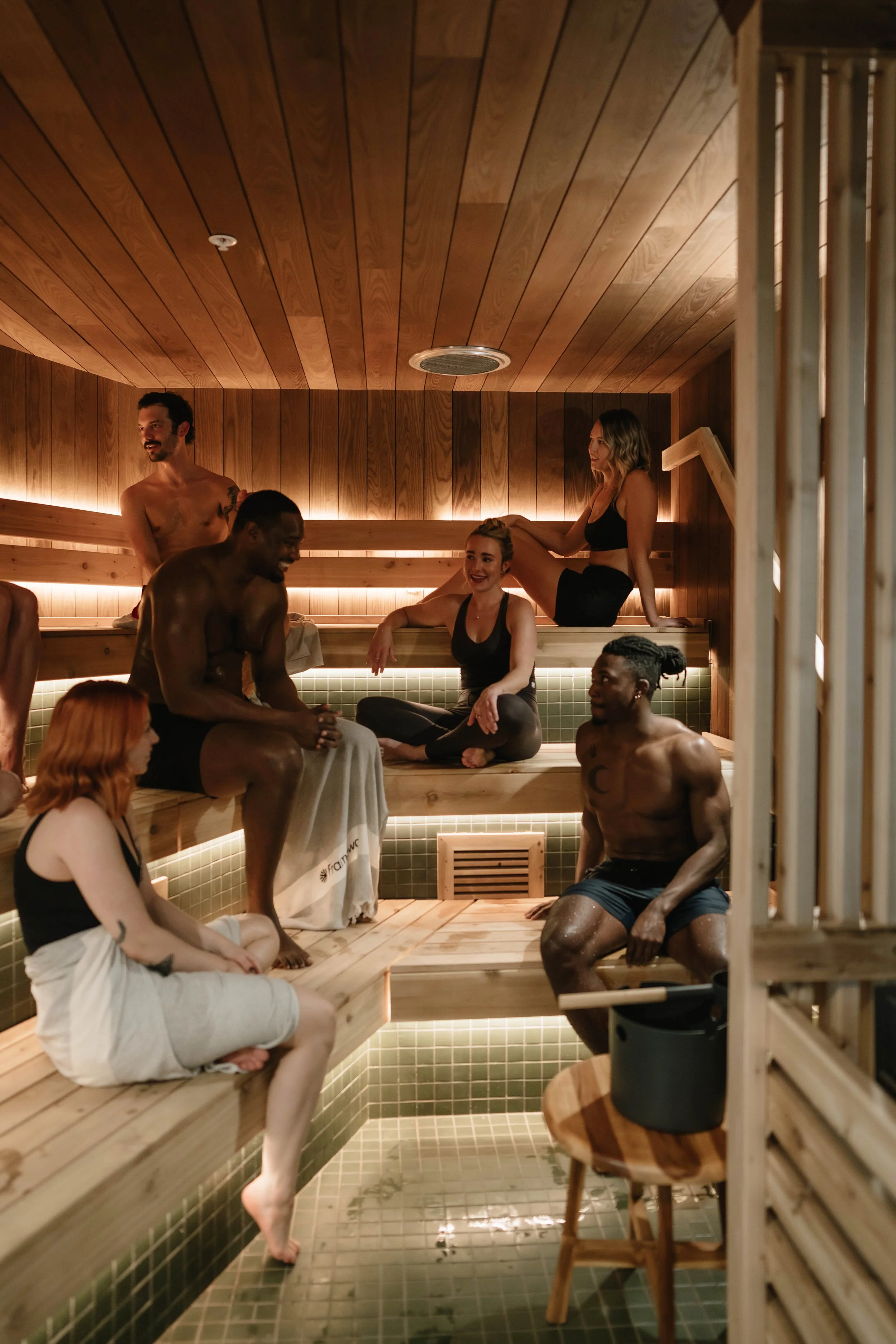 People enjoying a sauna, sitting on wooden benches, some with towels, in a room with wooden walls and ceiling, with warm lighting.