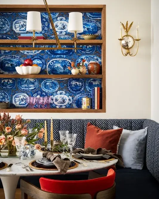 A dining area with a decorated table set with glassware, candles, and a floral arrangement, a cushioned bench with pillows, and a wall-mounted wooden shelf filled with decorative dishes and objects, with a blue patterned wallpaper background.