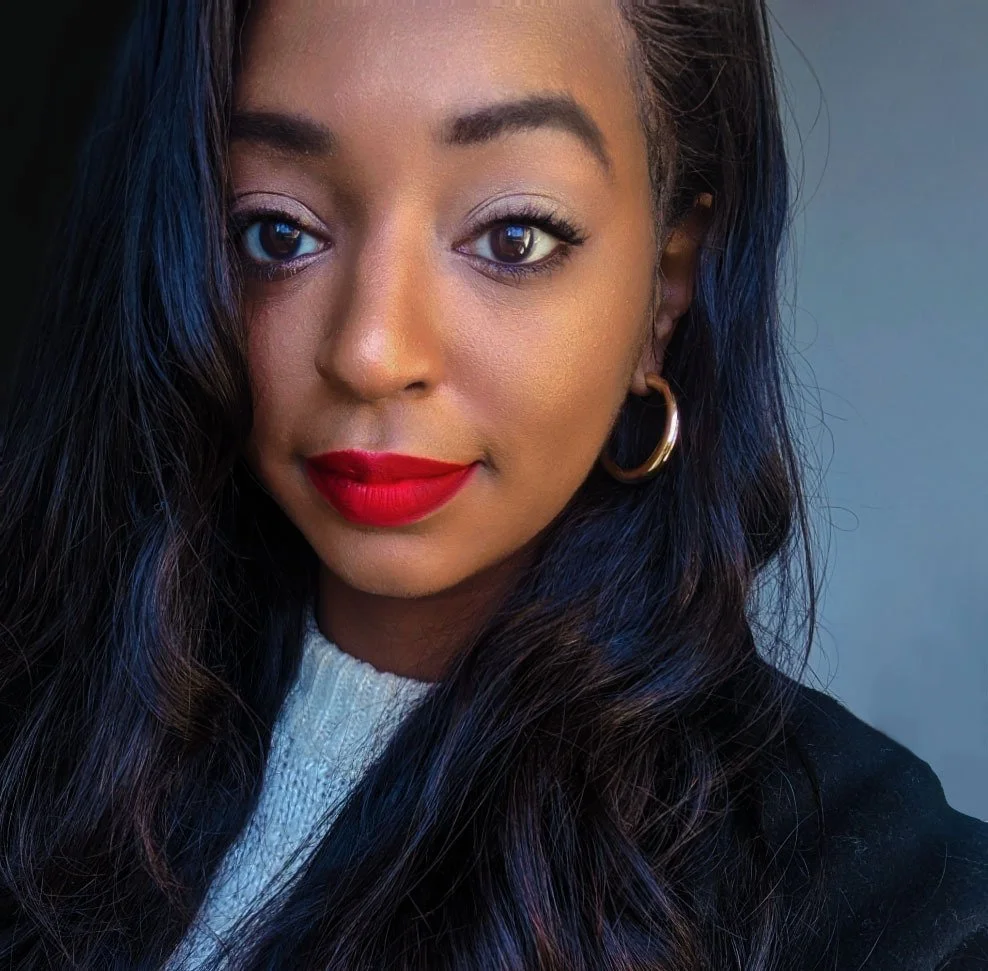 A young woman with dark wavy hair, red lipstick, and gold hoop earrings takes a close-up selfie against a neutral background.