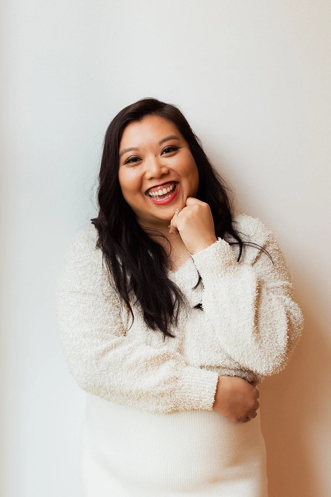 A smiling woman with long dark hair, wearing a cream cozy sweater, standing against a plain white wall.