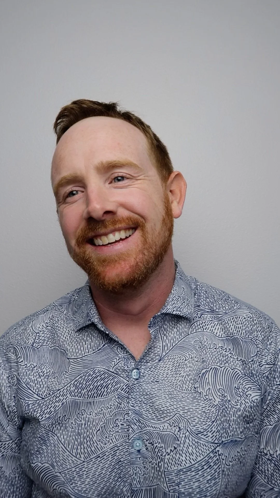 A smiling man with red hair and beard wearing a patterned button-up shirt, standing against a plain wall.