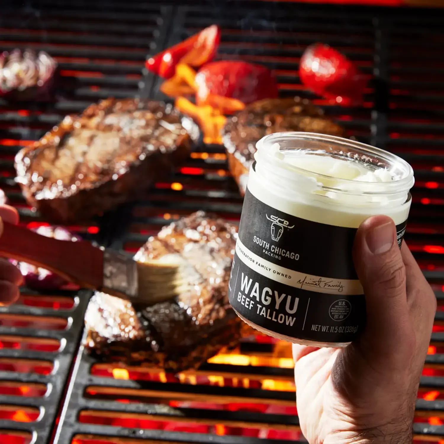 Person holding a container of Wagyu beef tallow near a barbecue grill with meat and vegetables cooking on it.
