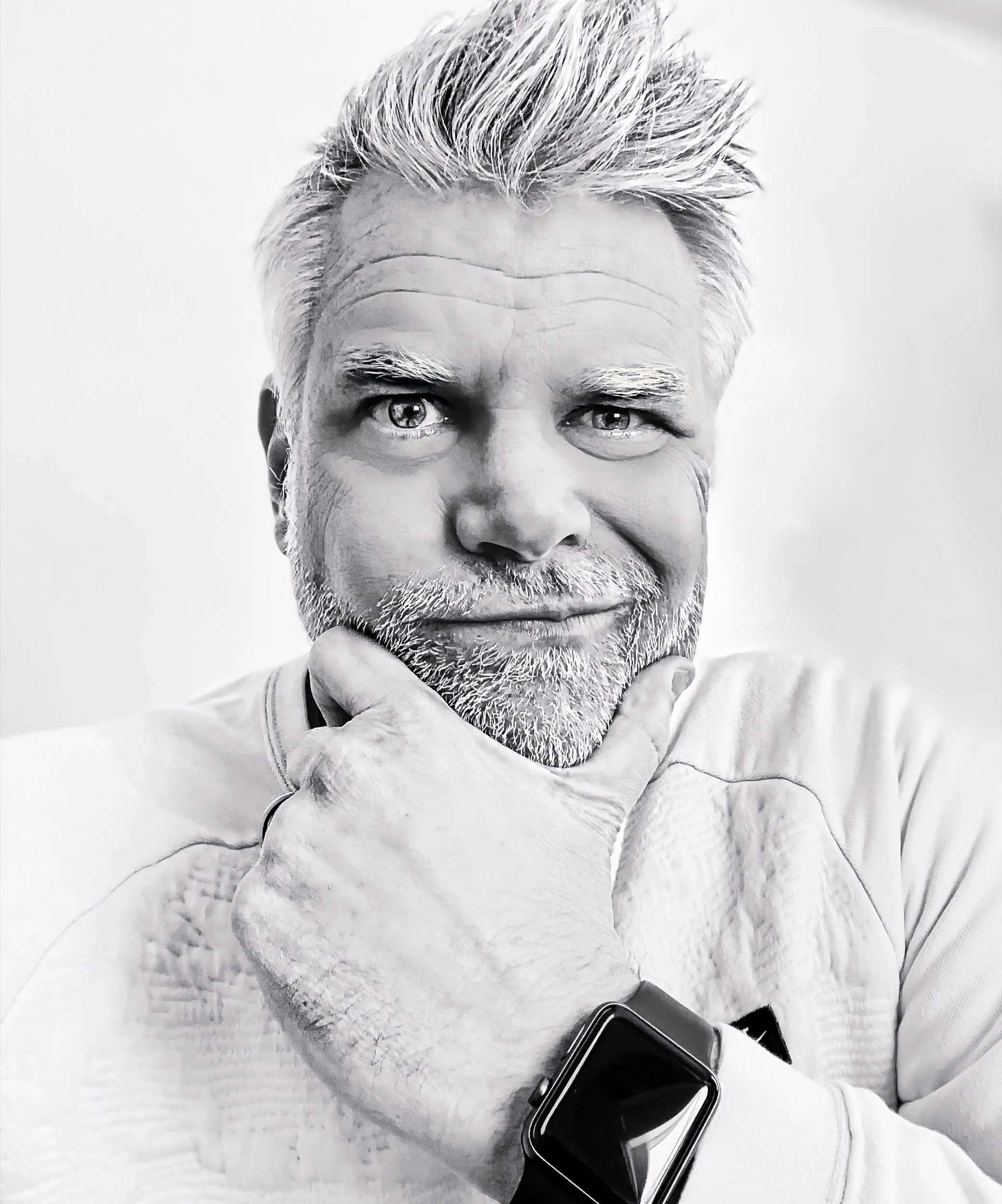 A man with white hair and beard, wearing a smartwatch, resting his chin on his hand, with a confident smile and looking directly at the camera.