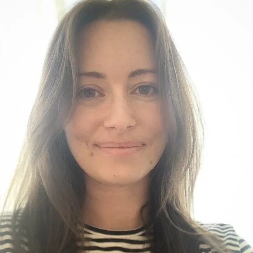 A young woman with light brown hair and a striped shirt smiling at the camera in a well-lit room.