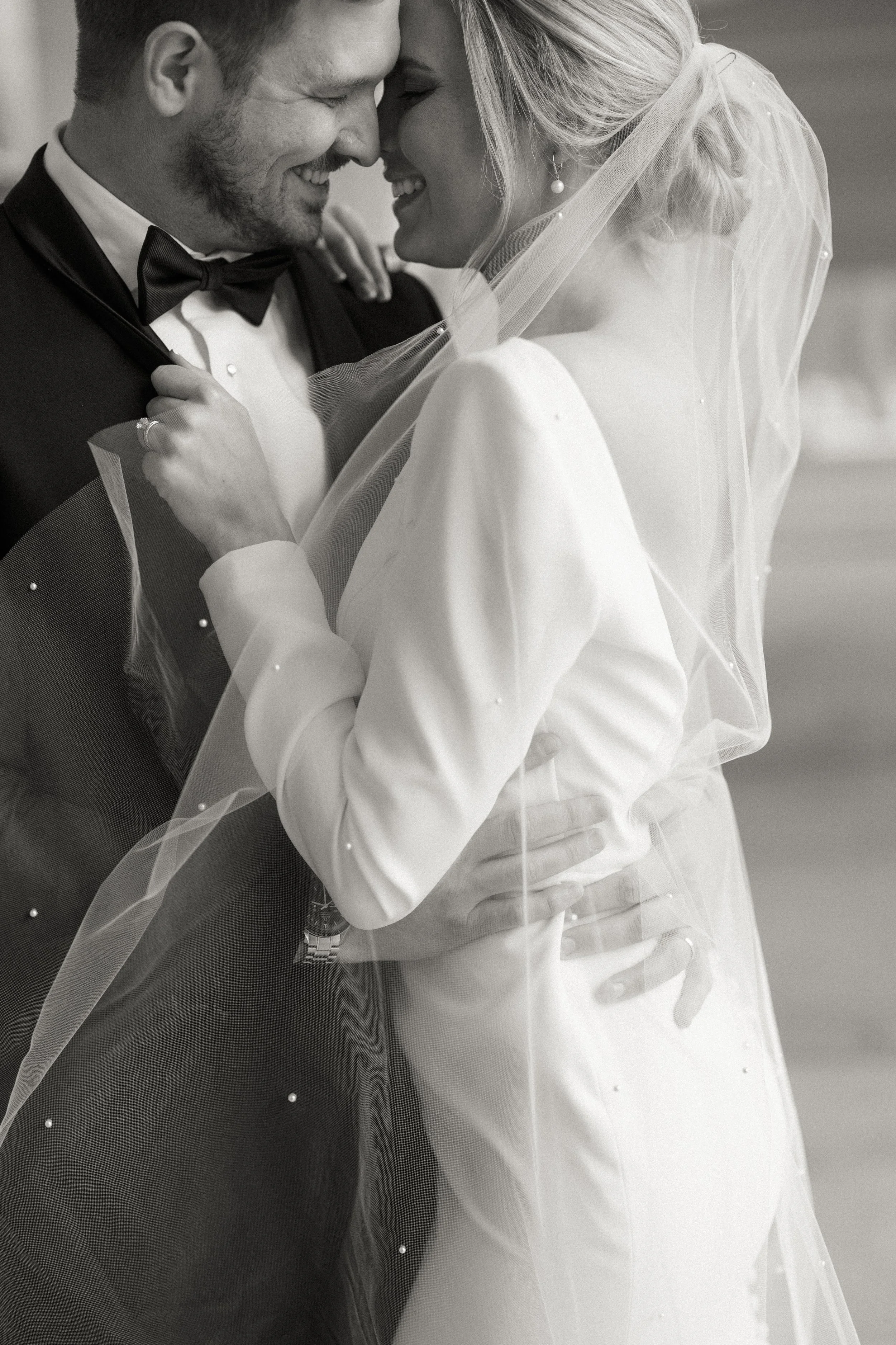 A black and white photo of a bride and groom sharing a close, intimate moment, smiling with their foreheads touching. The groom is wearing a tuxedo with a bow tie, and the bride is in a wedding dress with a veil.