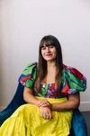 A woman with long brown hair and bangs wearing a colorful, patterned blouse with puffy sleeves and a yellow, lace skirt seated on a blue armchair against a white wall.