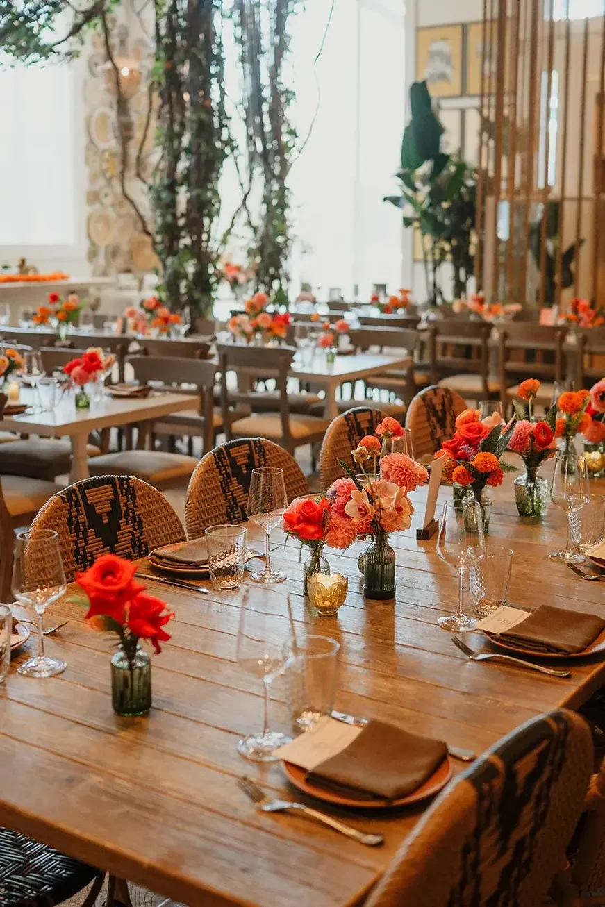 Indoor restaurant with wooden tables decorated with pink and orange flower centerpieces, set for a meal with glasses, plates, and napkins.