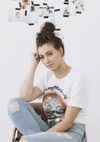 A woman with a top knot hairstyle sitting on a chair with torn jeans, wearing a white graphic t-shirt, looking at the camera with her hand resting on her head. In the background, a white wall with photos taped on it, including images of a woman by water and other scenes.