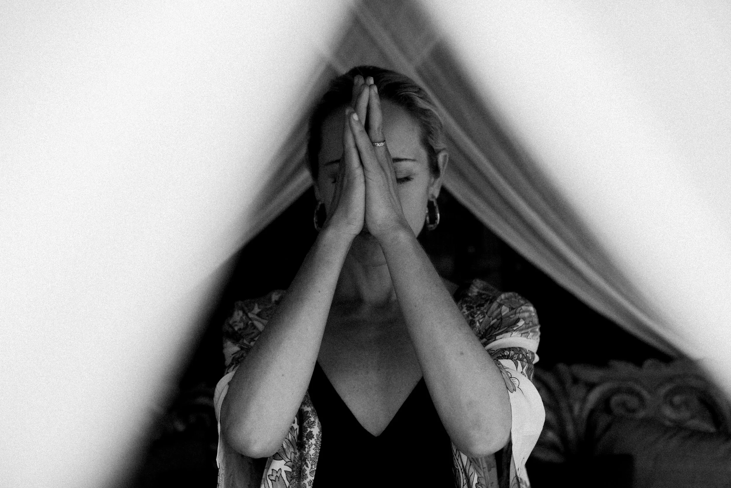 Black and white image of a woman with short hair, her hands pressed together in front of her face, eyes closed, in a contemplative pose, in a small, narrow space.