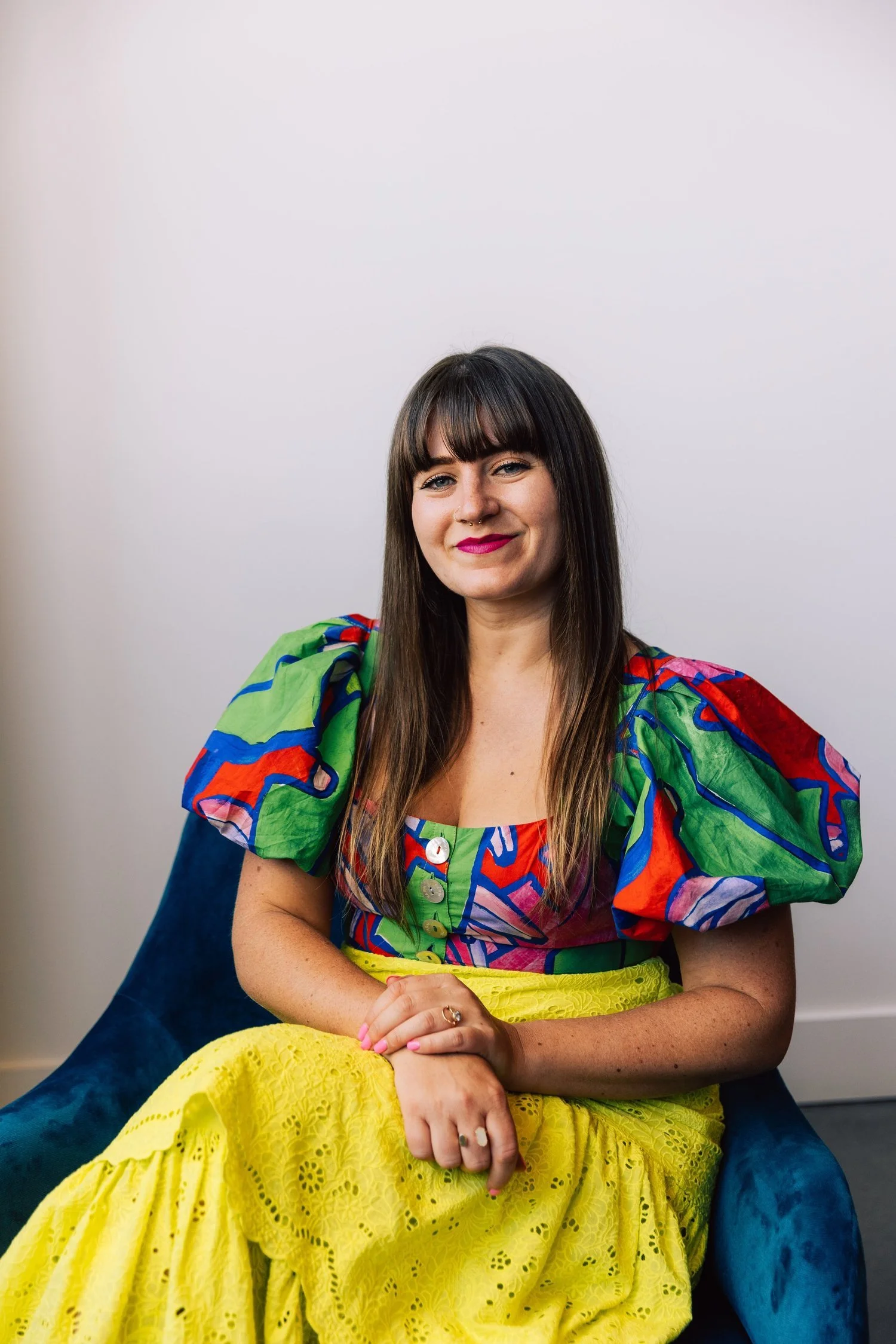 A woman with long brown hair and bangs wearing a colorful, patterned blouse with puffy sleeves and a yellow, lace skirt seated on a blue armchair against a white wall.