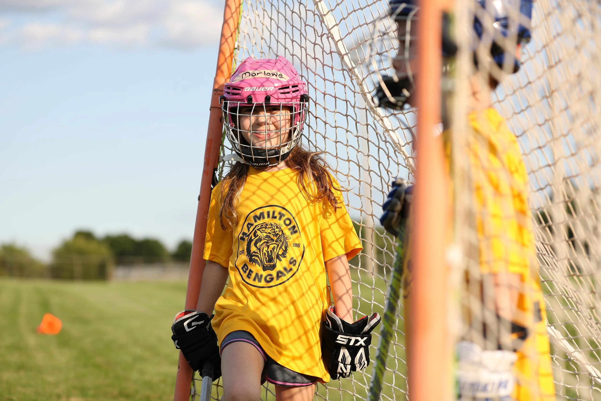 Young female goalie in yellow sports uniform with 'Clam Milton Bengals' logo, wearing a pink helmet with a face cage, standing behind a soccer goal net on a grassy field on a sunny day.