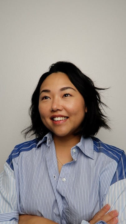 A woman with shoulder-length black hair, smiling, wearing a blue and white striped shirt, standing against a plain wall.