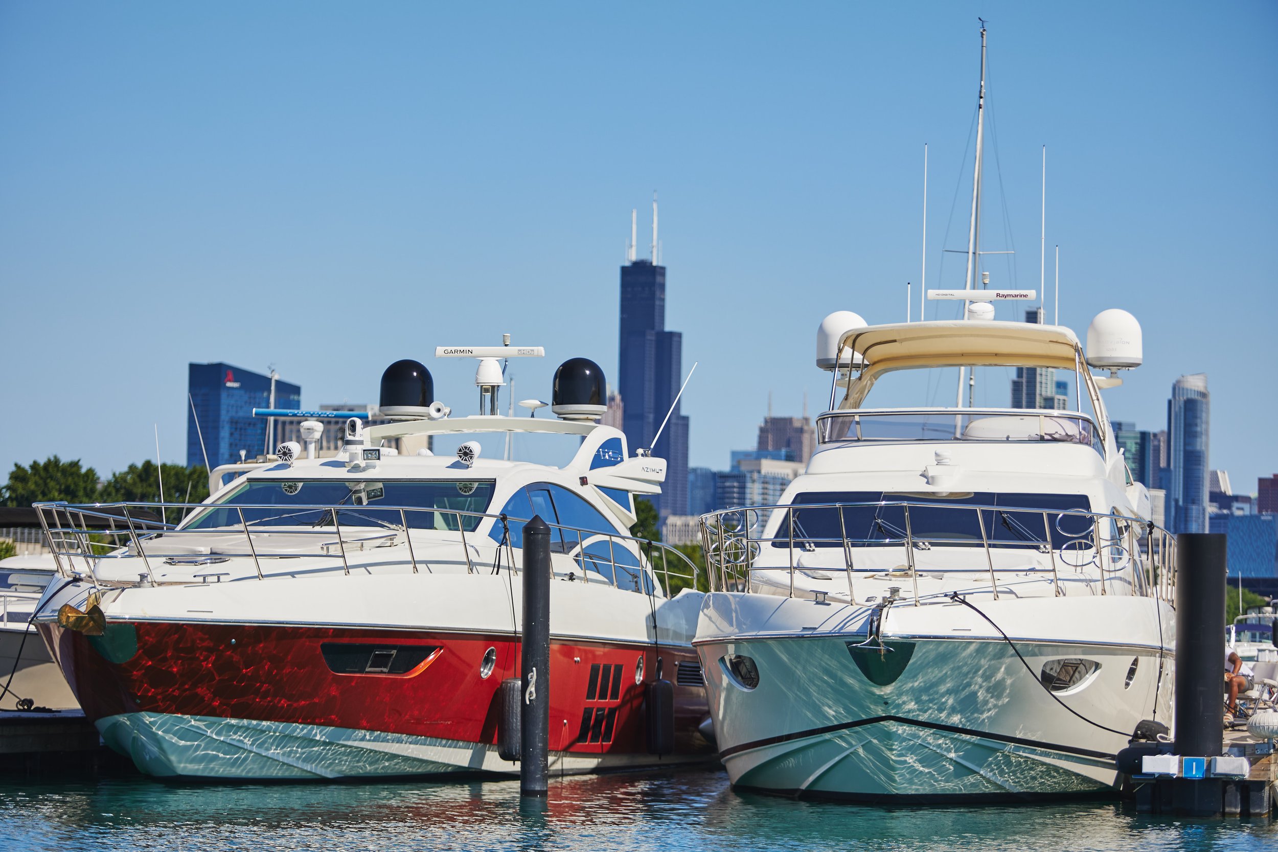 Several luxury yachts docked at a marina with city skyscrapers in the background on a clear, sunny day.
