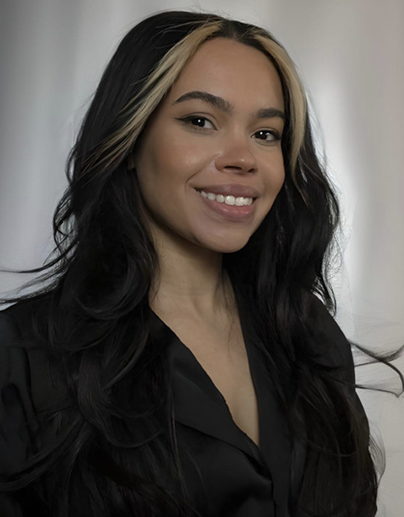 A woman with long dark hair featuring blonde highlights, smiling and wearing a black shirt, standing in front of a plain wall.