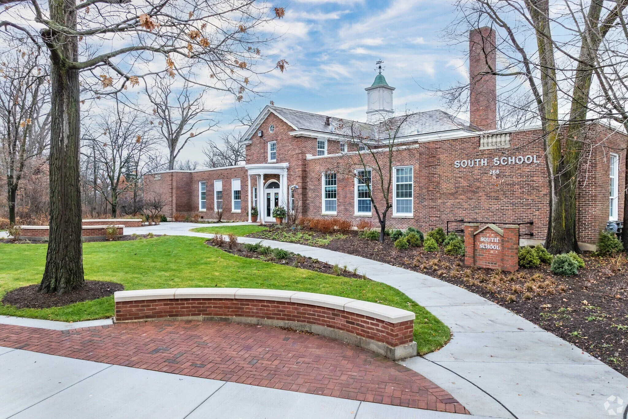 A brick school building labeled 'South School' with a tall brick chimney and a small tower with a weather vane on top. The surrounding yard has green grass, leafless trees, and a curved brick and concrete walkway.