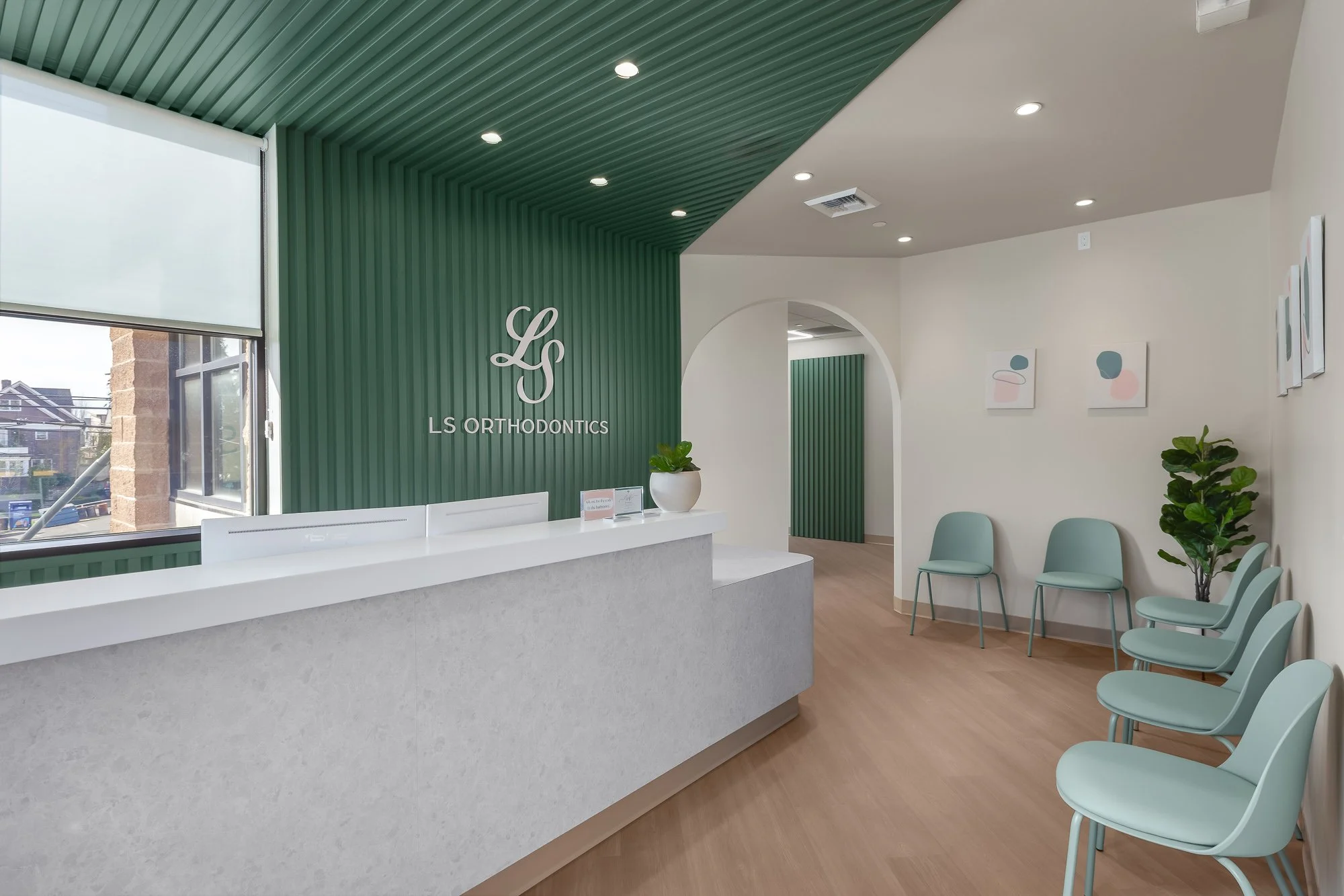 Reception area of LS Orthodontics with a white counter, green accent wall with the clinic's logo, framed abstract art on white walls, and light green chairs for patients, with indoor plants and a large window letting in natural light.
