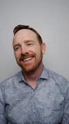 A smiling man with red hair and beard wearing a patterned button-up shirt, standing against a plain wall.