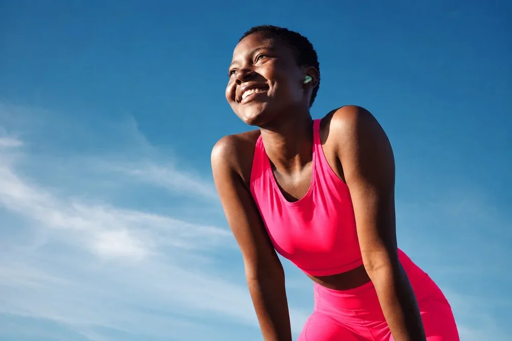 A woman smiling and looking away while leaning forward outdoors in workout attire against a bright blue sky.