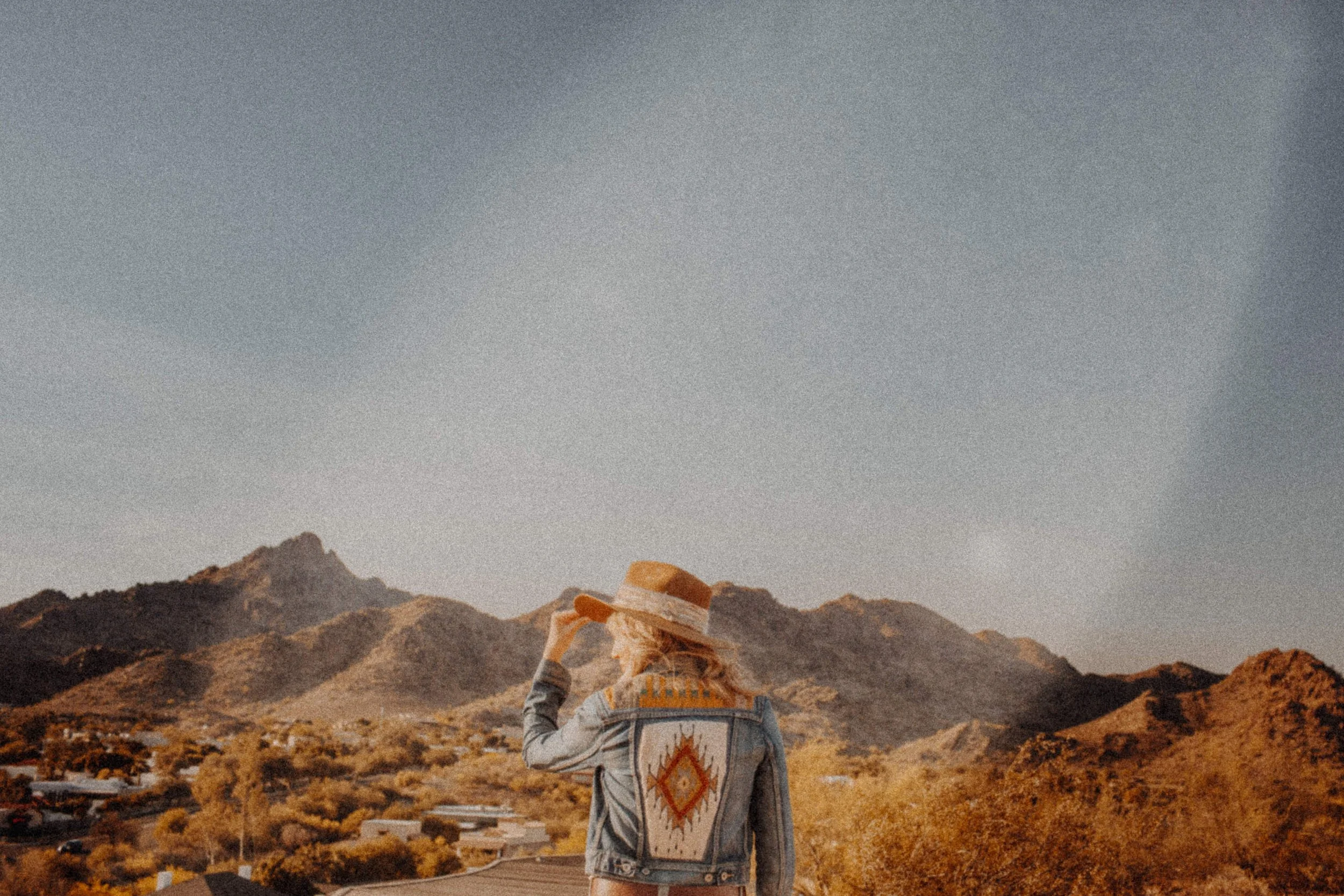 A person with a hat and a distinctive jacket walking through a desert landscape with mountains in the background under a clear sky.