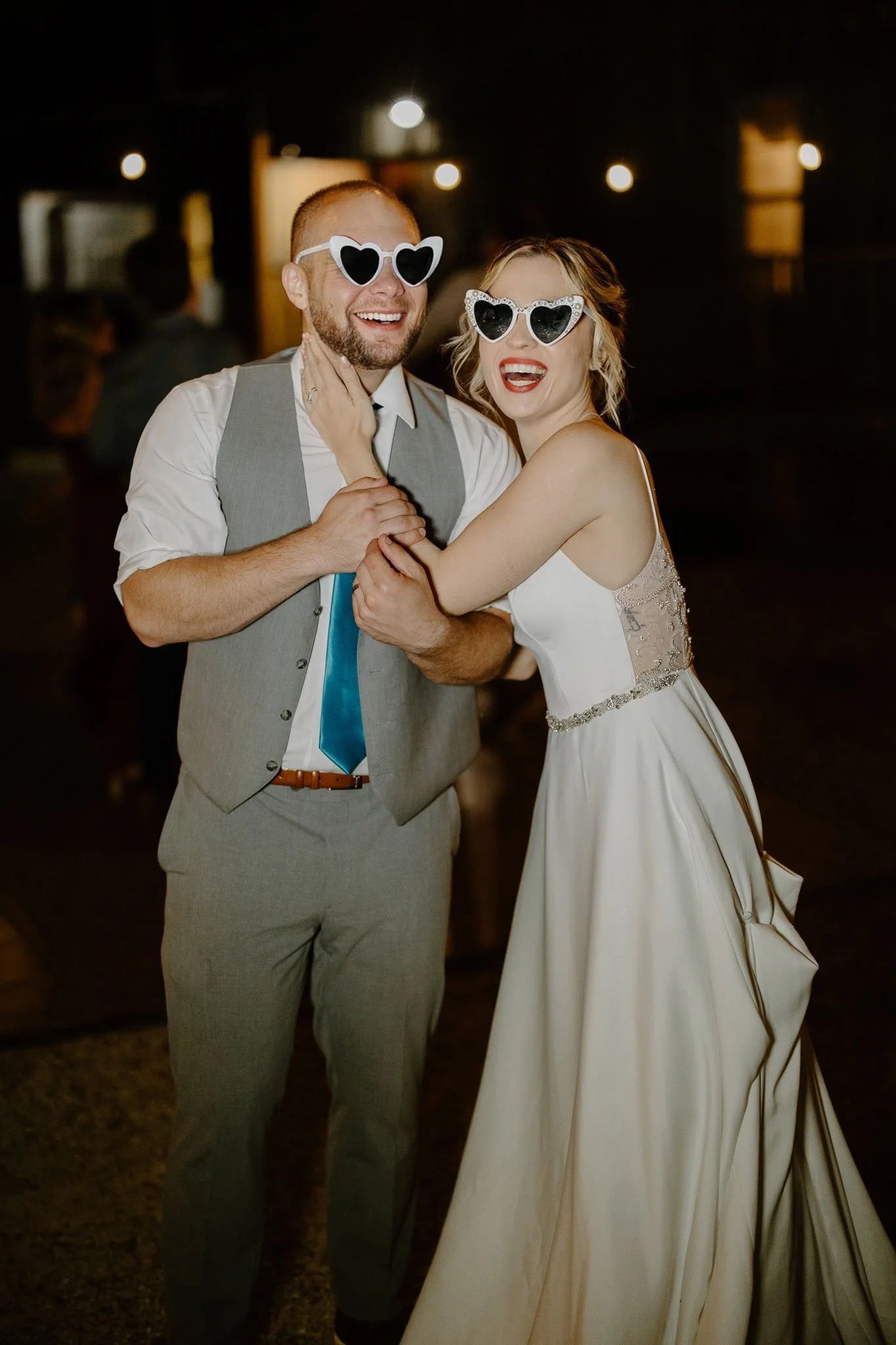 A happy bride and groom wearing heart-shaped sunglasses celebrate at their wedding reception. They are smiling and holding hands.