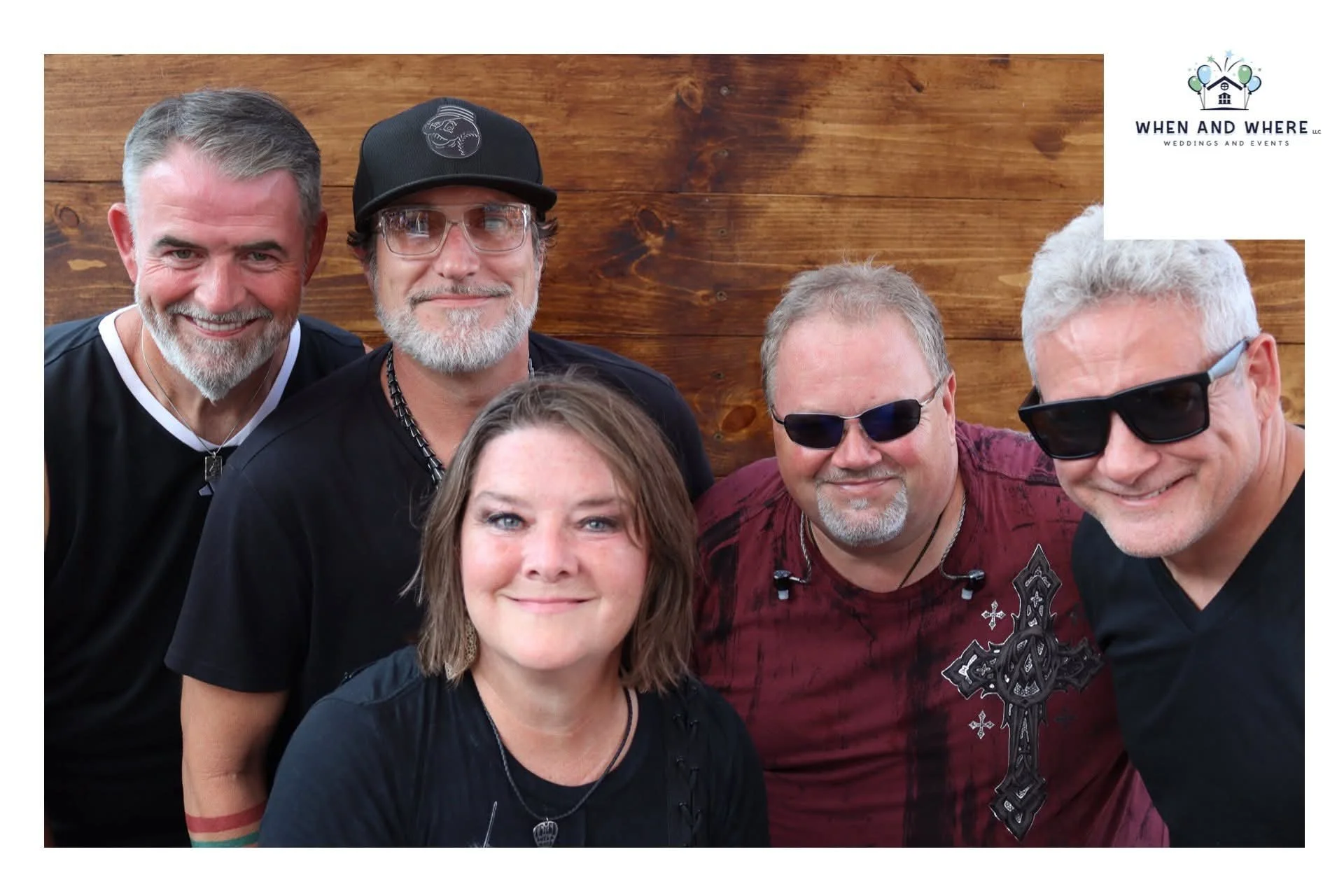 A group of five cheerful people, four men and one woman, posing indoors in front of a wooden wall. They are wearing casual clothing and sunglasses, with the woman and one of the men smiling at the camera.