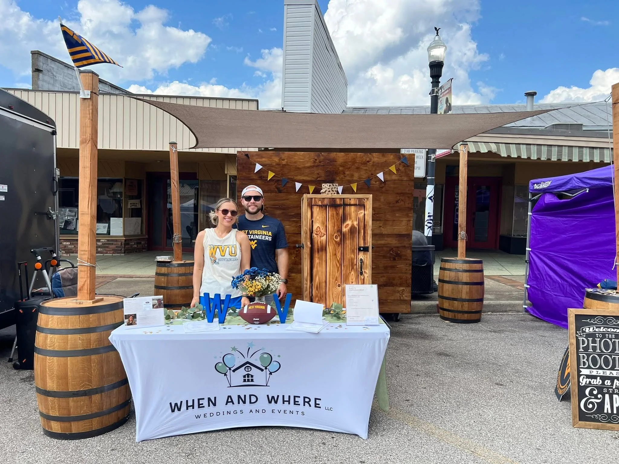 A photo of a man and woman standing behind a table at an event booth for wedding and event planning. The table is covered with a white tablecloth that has the logo of 'When and Where Weddings and Events LLC'. The booth is decorated with a wooden back