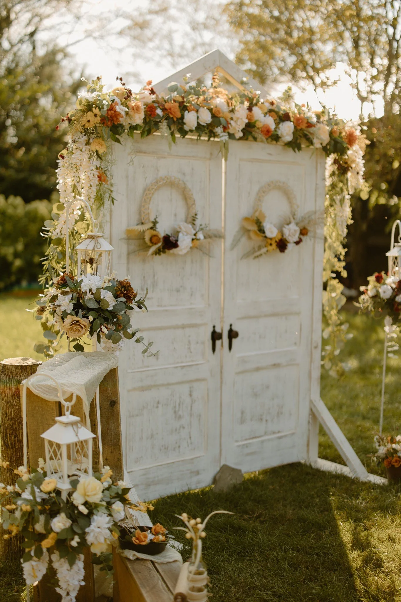 Decorative outdoor wedding backdrop with white wooden doors, floral arrangements, lanterns, and greenery.