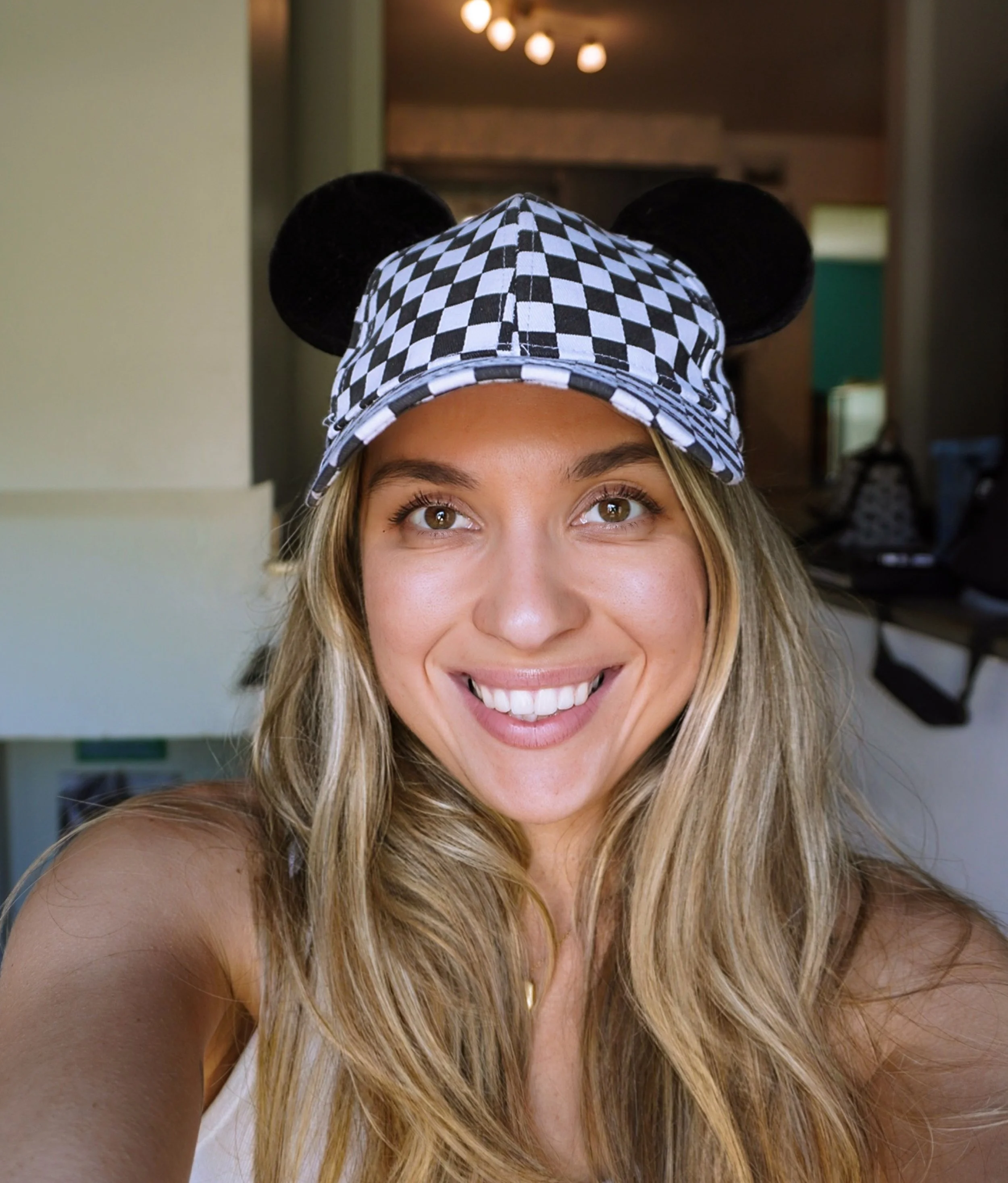 A woman taking a selfie, wearing a checkered hat with black Mickey Mouse ears, smiling brightly.
