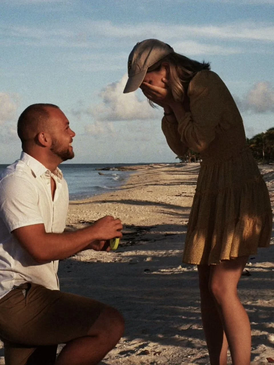 A man kneeling on the beach, proposing to a woman who is covering her mouth in surprise, both smiling, in front of the ocean during sunset.