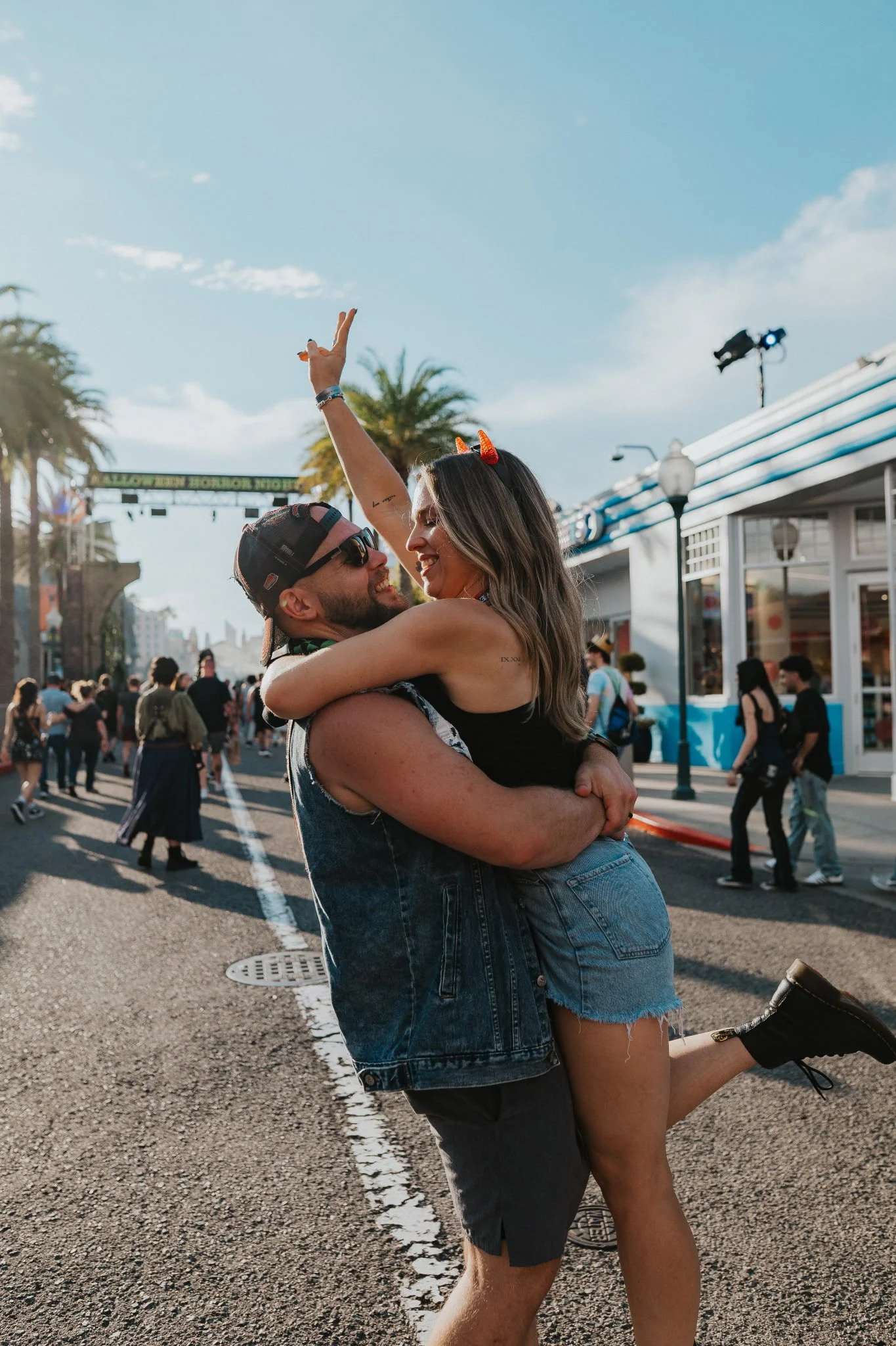 A couple enjoying an amusement park on a sunny day, with the man lifting the woman while they smile and celebrate.