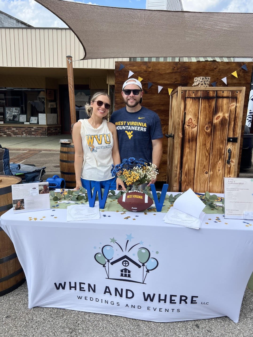 Two people standing behind a table with promotional materials for weddings and events, dressed in West Virginia Mountaineers apparel, at an outdoor event.