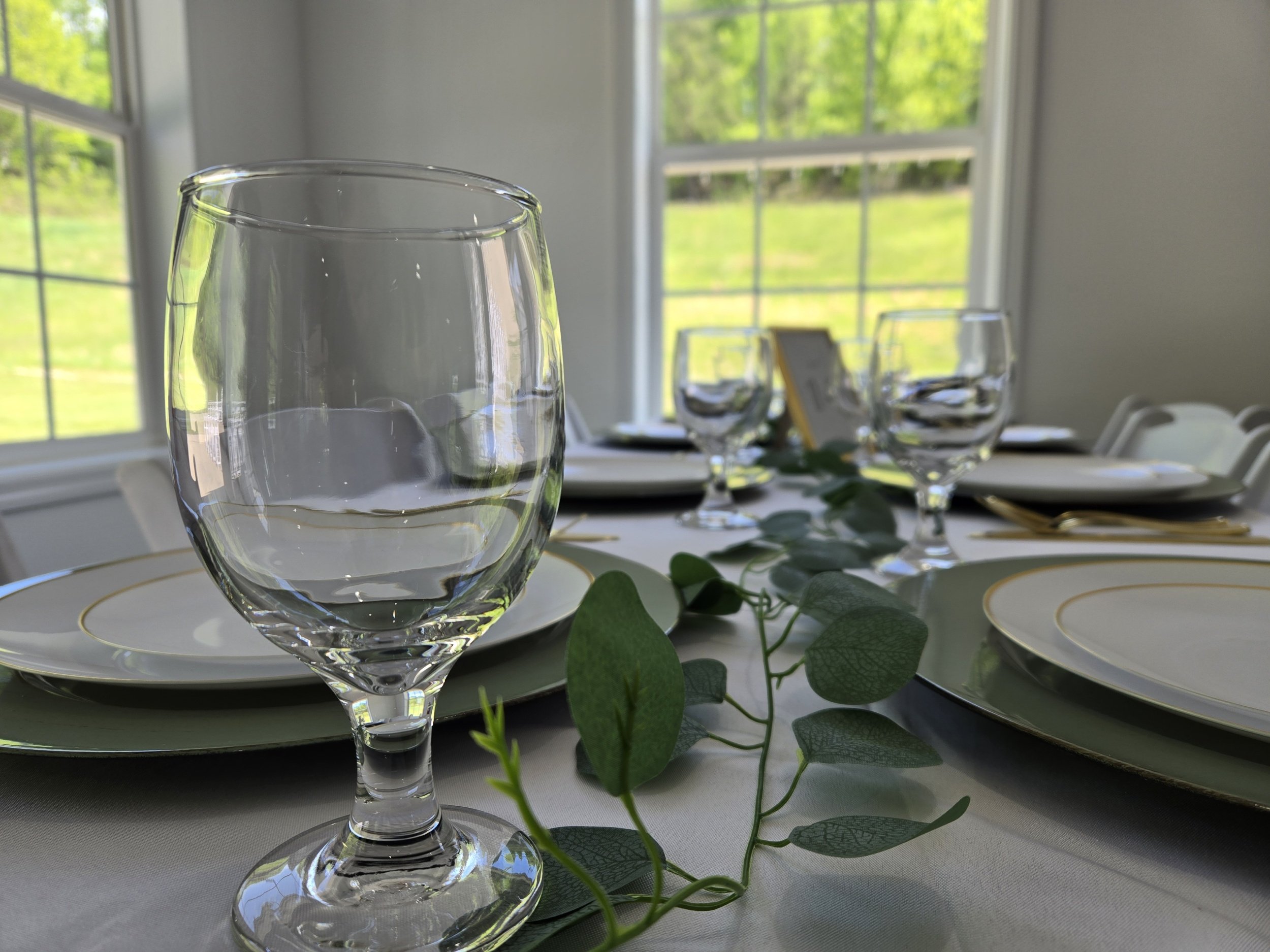 A dining table set with clear wine glasses, white plates with gold trim, green napkins, and gold utensils, decorated with green leaves, in a room with large windows showing a green outdoor landscape.