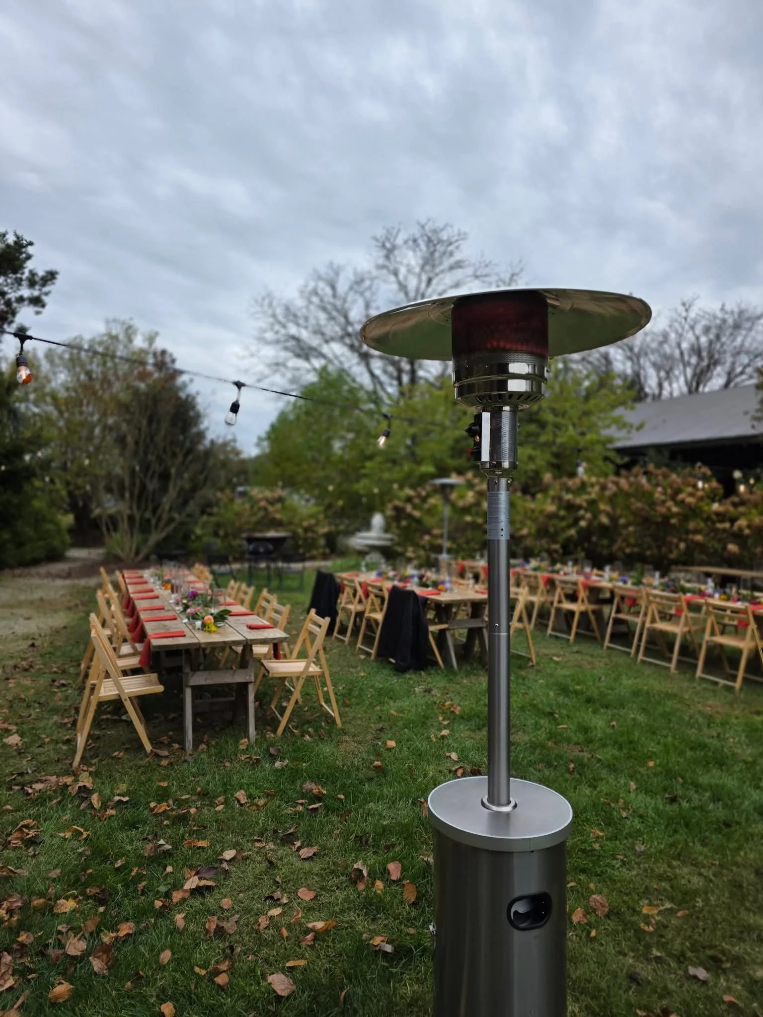 Outdoor garden party setup with long tables decorated with flowers and red napkins, surrounded by wooden chairs, under a cloudy sky.
