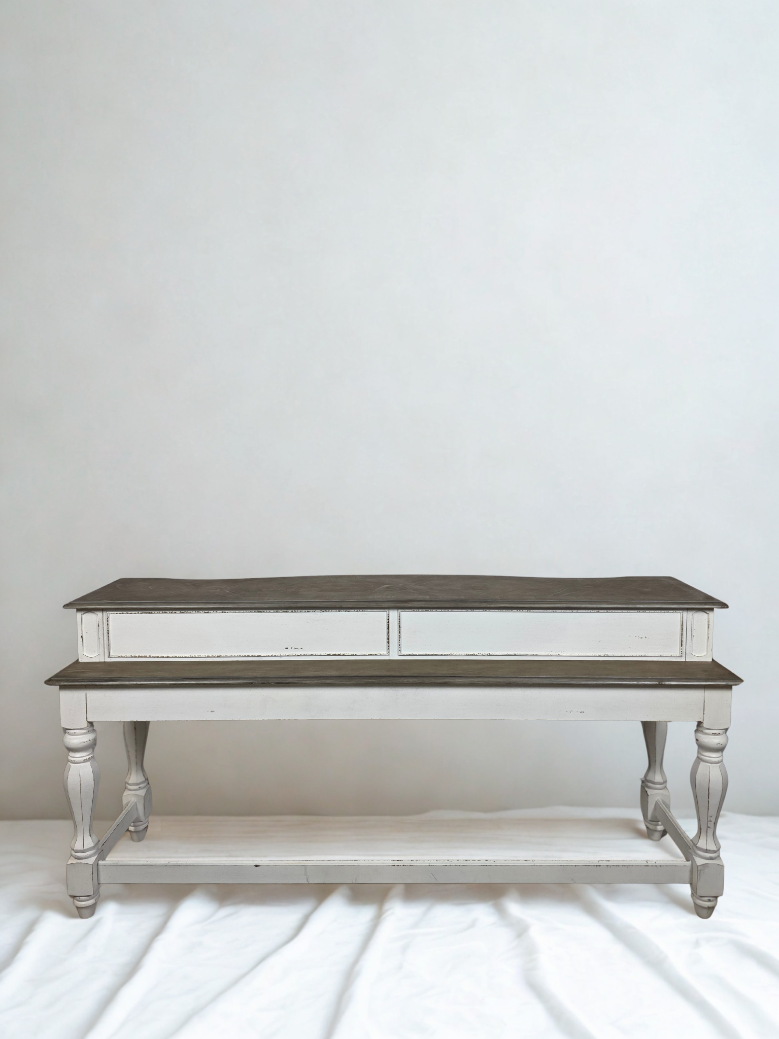 A vintage-style wooden coffee table with a two-tone finish, featuring a darker top surface and lower shelf, with light-colored, turned legs and side panels, set against a plain white wall.
