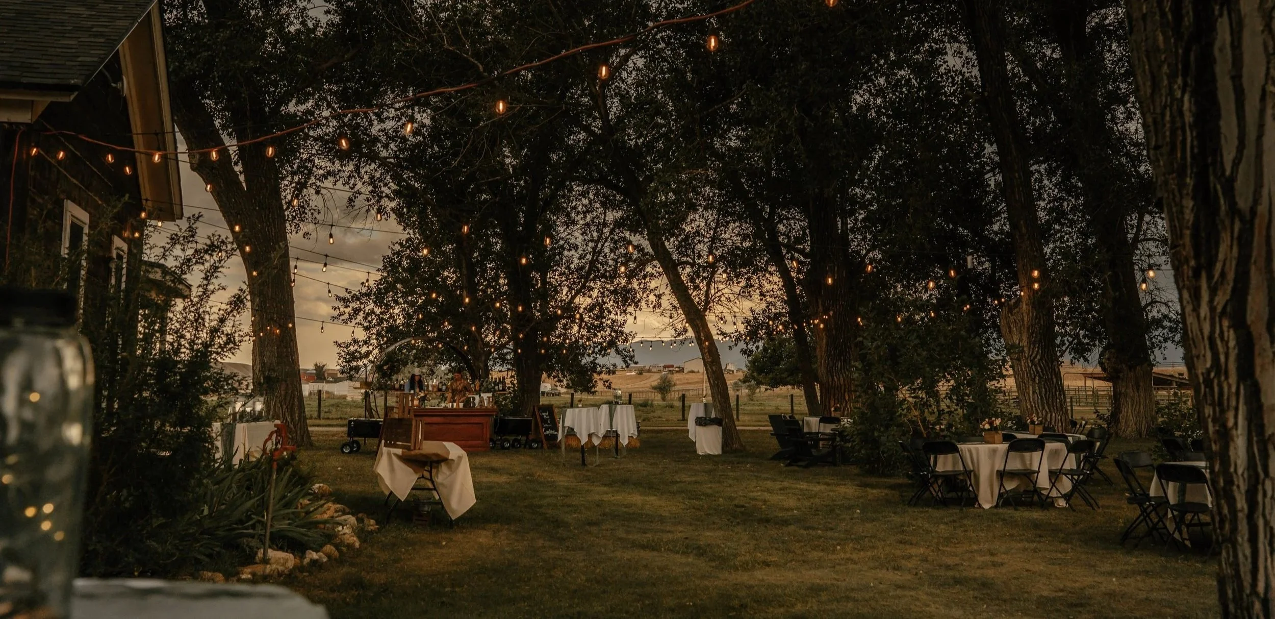 Outdoor evening setting with string lights hanging between trees, tables with white tablecloths, and chairs, possibly indicating a party or event, in a rural or countryside area with trees and open fields in the background.
