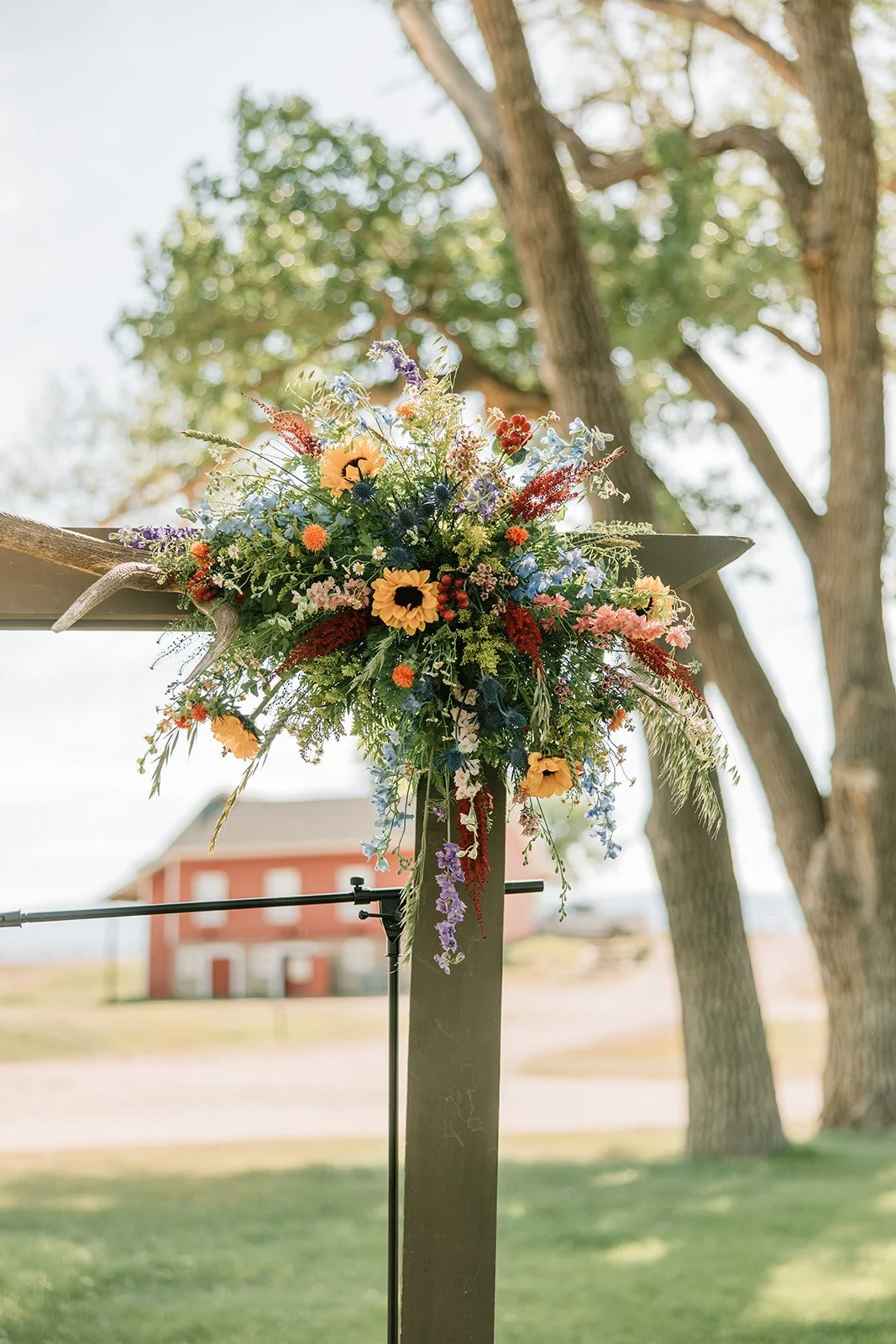 Colorful floral arrangement with sunflowers, blue, purple, red, and white flowers attached to a black post outdoors, with trees and a red building in the background.