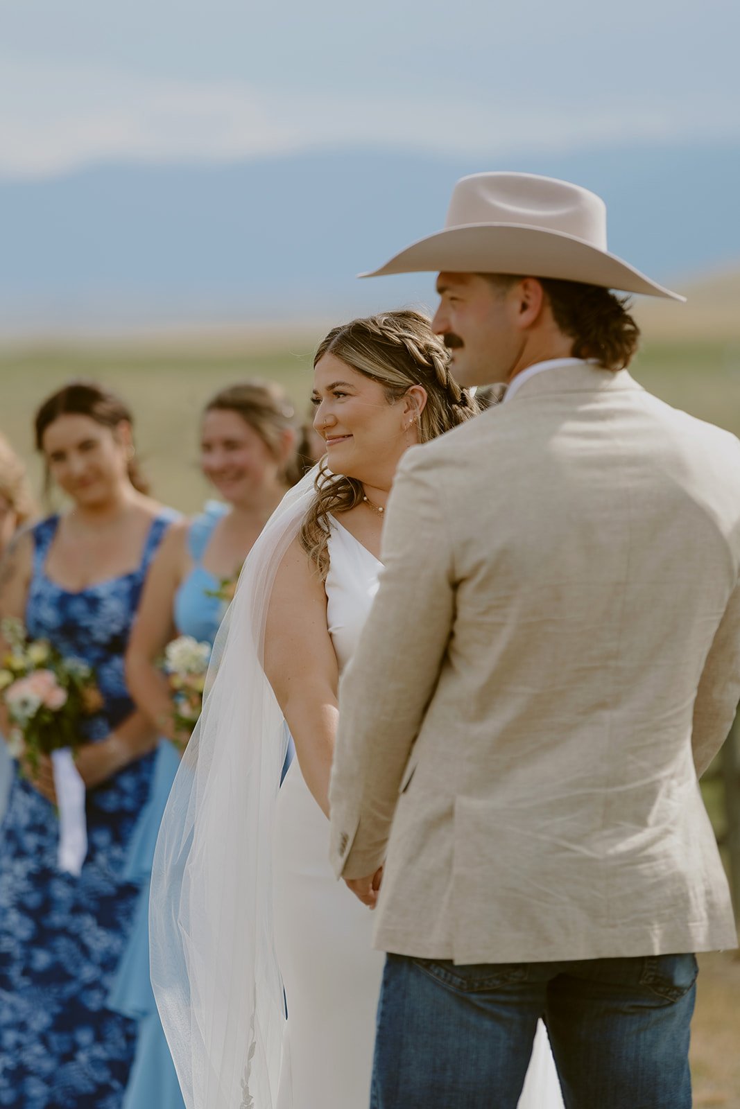 A wedding ceremony outdoors with a bride and groom holding hands, surrounded by bridesmaids in blue dresses and floral bouquets, with a scenic mountain landscape in the background.