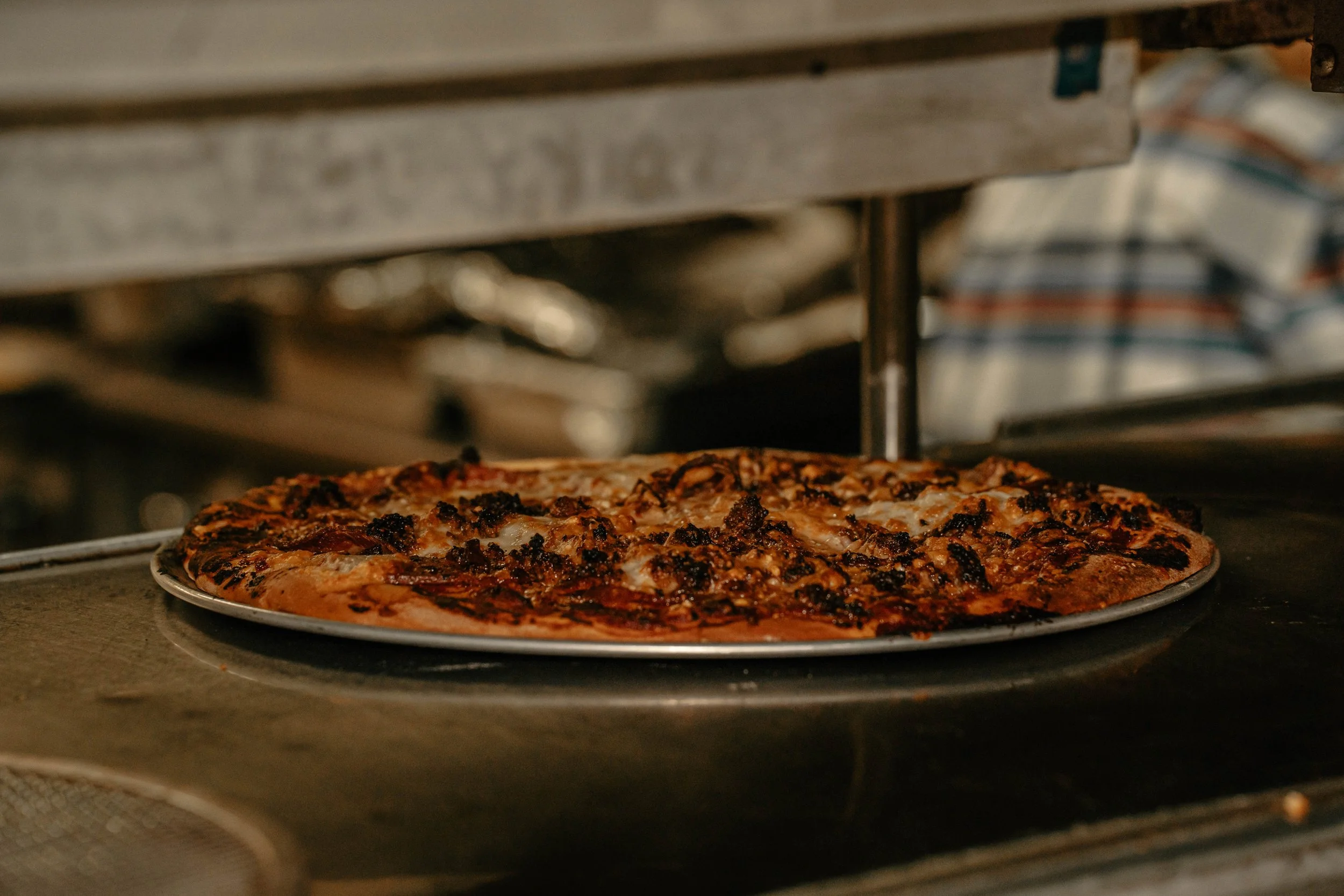 Freshly baked pizza with cheese, tomato sauce, and dark toppings on a metal tray in a pizzeria kitchen.