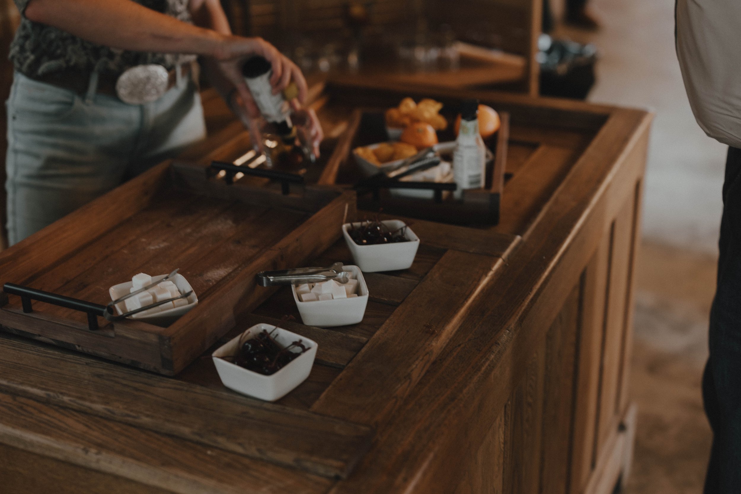 A bar cart with orange and yellow fruits, bottles of bitters, and other cocktail ingredients. A person in a striped shirt is reaching for a jar in the background.