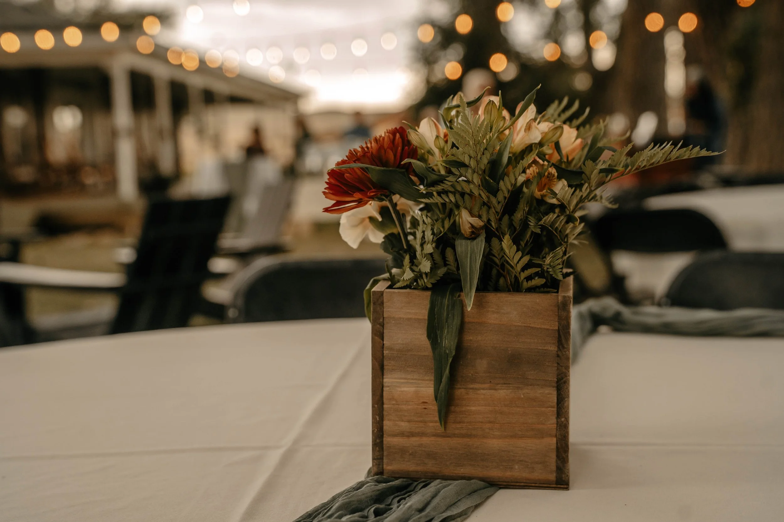 A centerpiece of flowers in a wooden box on a table with a cream tablecloth at an outdoor event, with string lights and blurred people in the background.