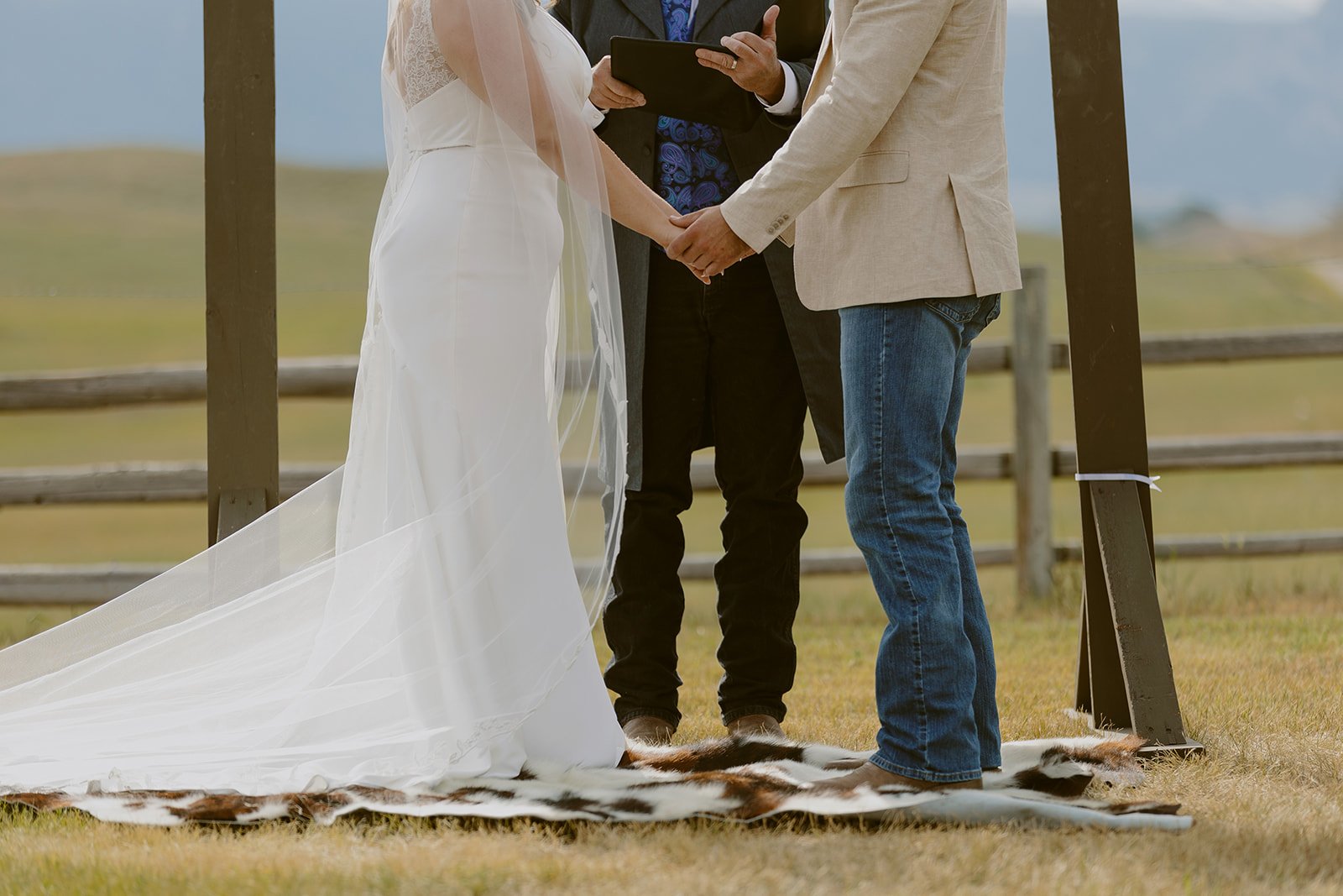 A couple is getting married outdoors, holding hands, with an officiant holding a tablet in front of them. They are standing on a cowhide rug, with a wooden arch and fields in the background.