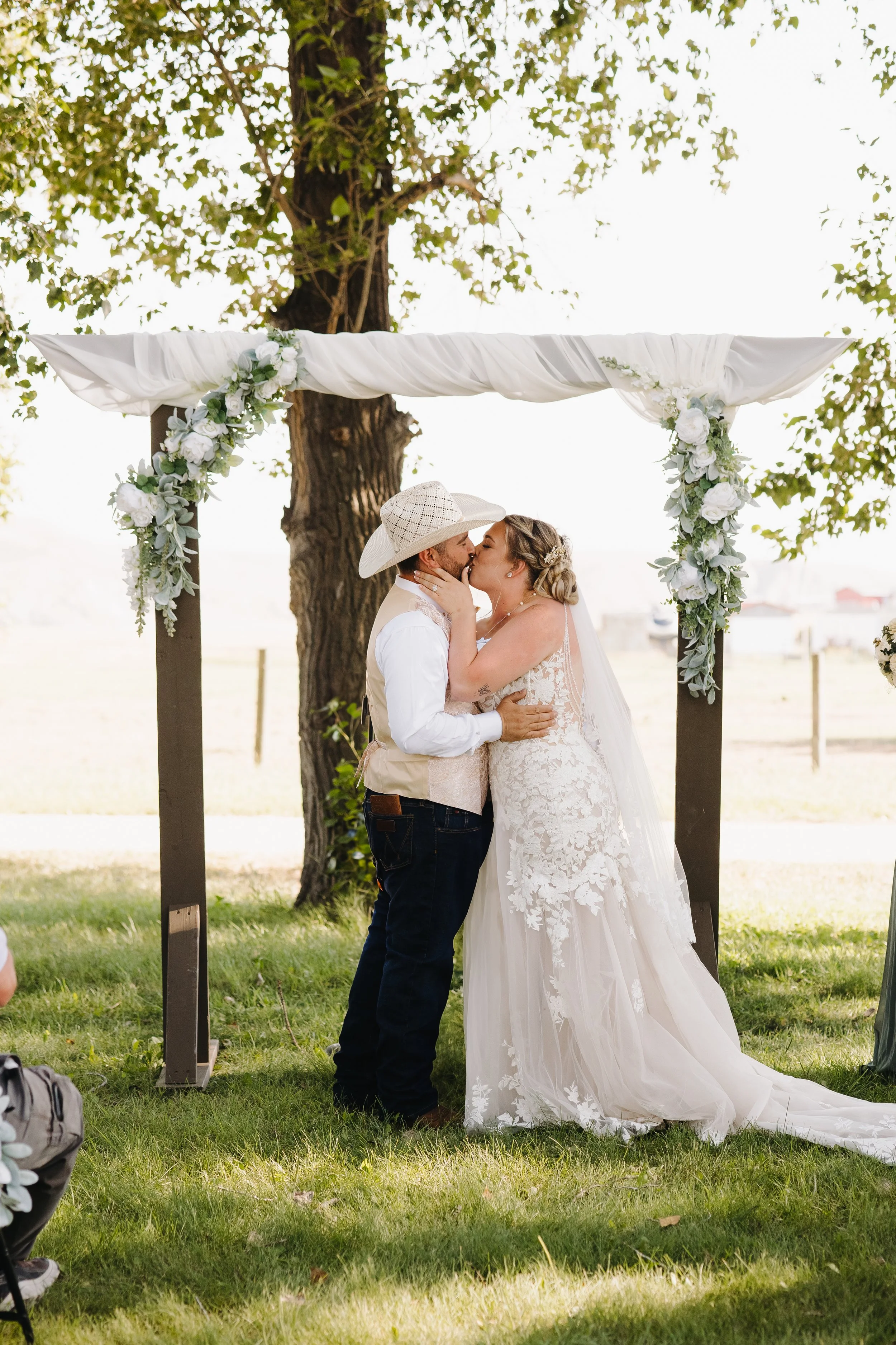 A couple kissing at their outdoor wedding ceremony, under a wooden arch decorated with white flowers and green foliage. The bride wears a lace wedding dress and veil, while the groom wears a cowboy hat and casual attire.