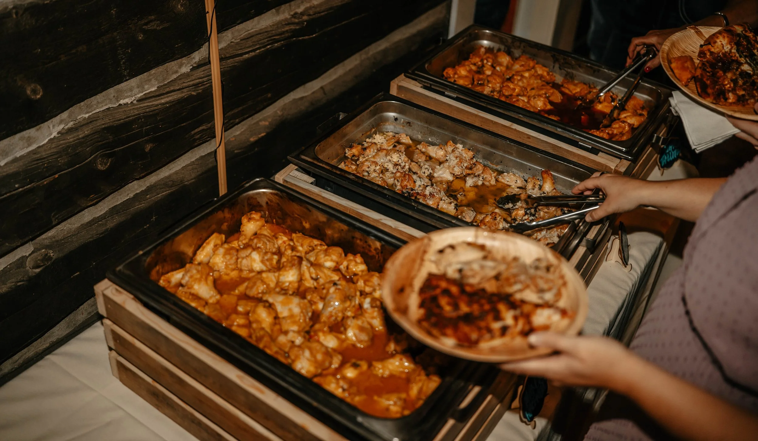 Buffet with trays of cooked chicken dishes, with a person serving food onto a plate.