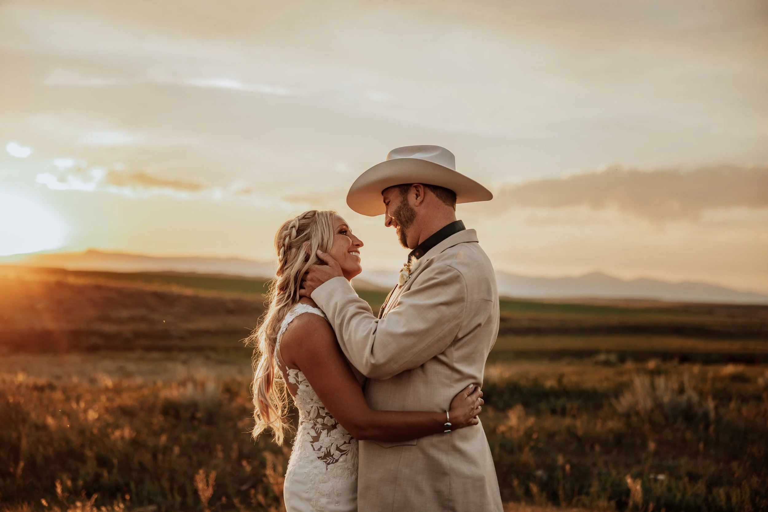 A bride and groom standing close together outdoors at sunset, gazing into each other's eyes with a scenic landscape in the background.