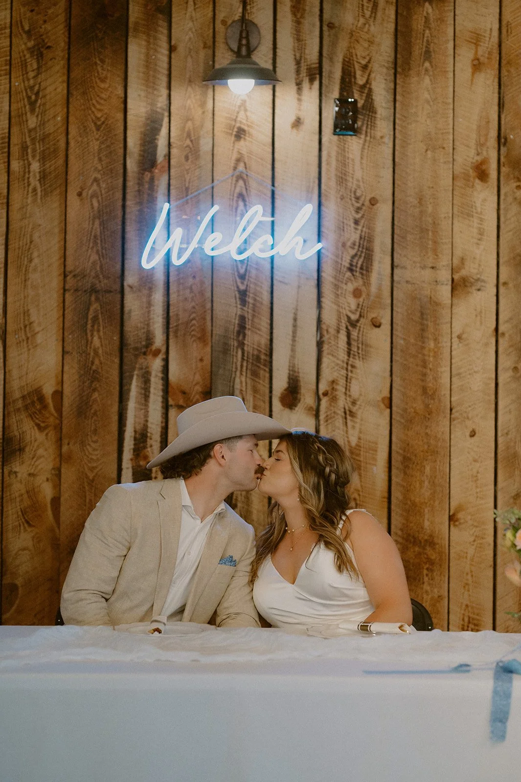 A couple kissing at a wedding reception, with a neon sign saying 'Welsh' on a wood-paneled wall behind them.